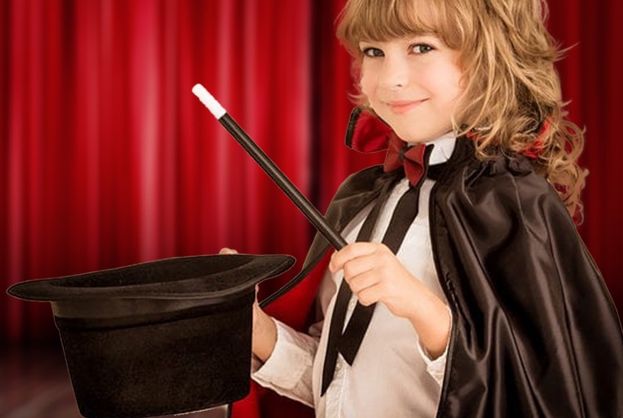 A child wearing a magician's cape, holding a wand and top hat in front of red curtains.