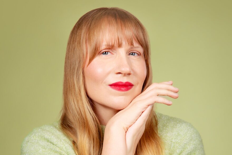 A woman with long strawberry blonde hair holds her hands up to her face and smiles slightly, her lips bright red. 