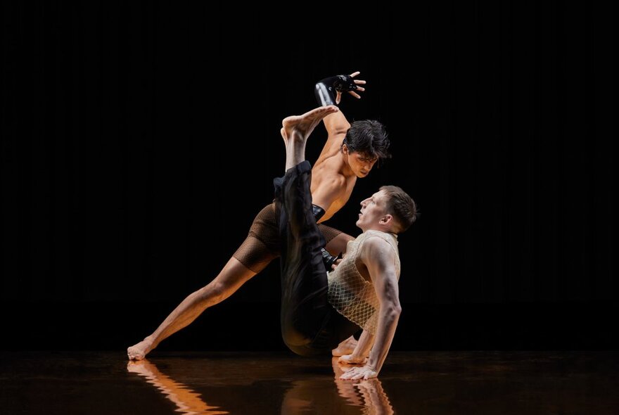 Two dancers in a closely held pose against a black background, one standing and leaning over the top of the other dancer who is sitting on the floor, their legs raised and their arms pressed down on the ground supporting their torso.