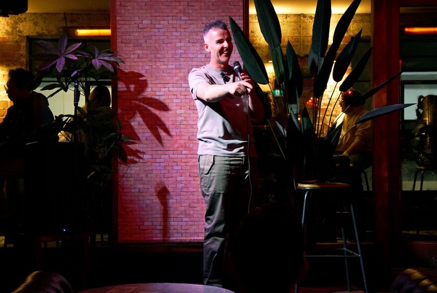 Comedian holding a microphone, standing under a spotlight in a bar with plants and bar stools.