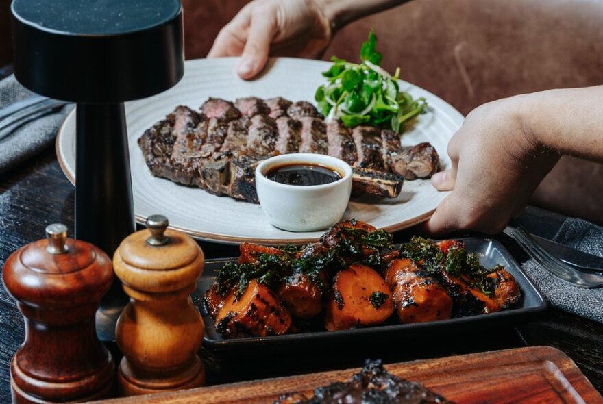 Hands placing a dish of steak onto a restaurant dining table next to a side dish of roasted vegetables.