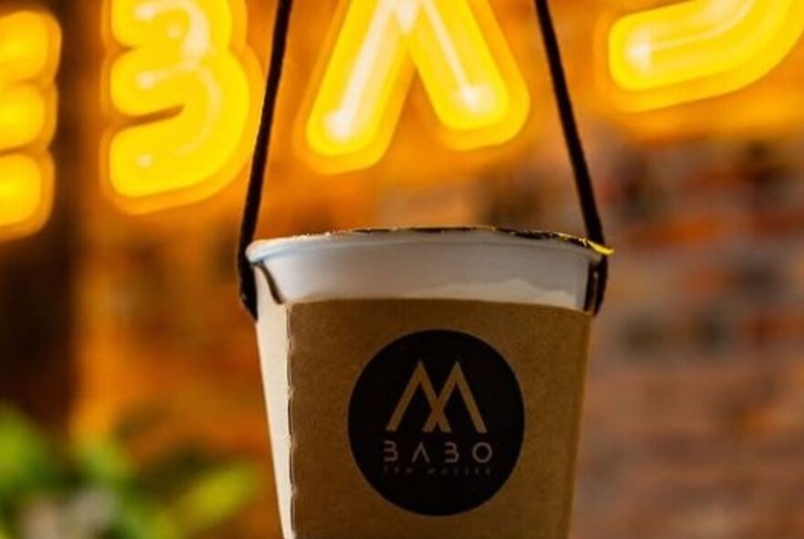 An out of sight hand holding a takeaway drink bubble tea with a string handle, with yellow neon cafe signage in the background.