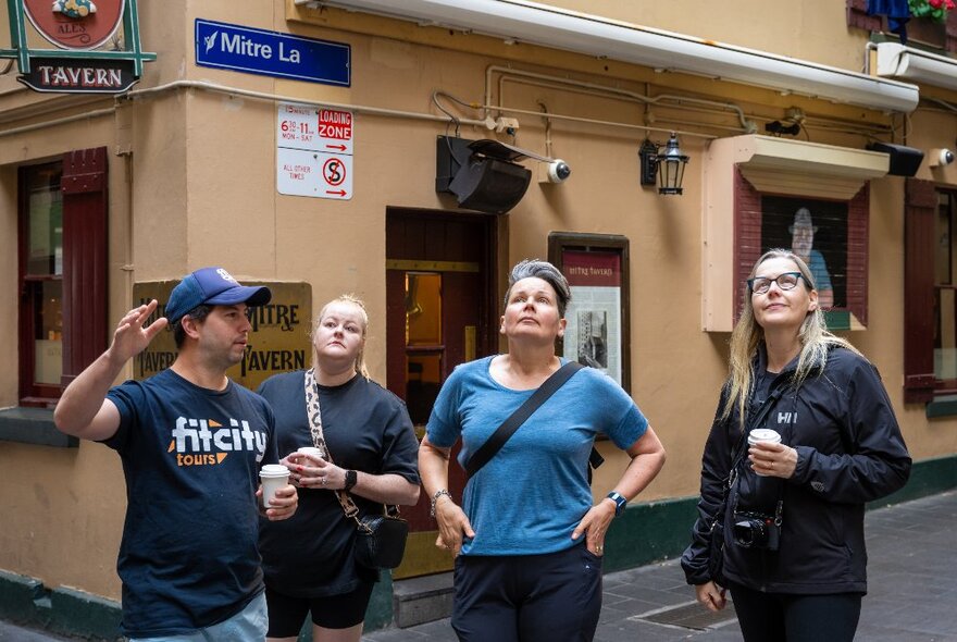 Three people on a walking tour with a Fit City tour guide, all looking up, the peach-coloured Mitre Tavern behind them.