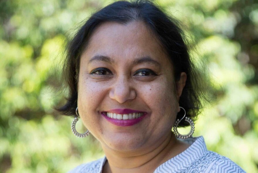 Close-up of the face of Mridula Nath Chakraborty, a smiling woman with short dark hair and hoop earrings, standing outside with greenery in the background.