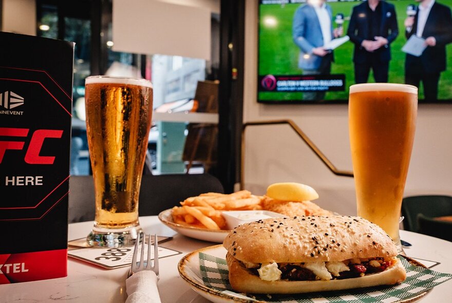 A table set with pub-style food and two glasses of beer with a football match on a screen in the background. 