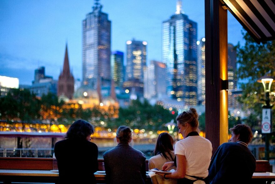 Customers and staff at restaurant tables overlooking the river and city buildings at dusk.