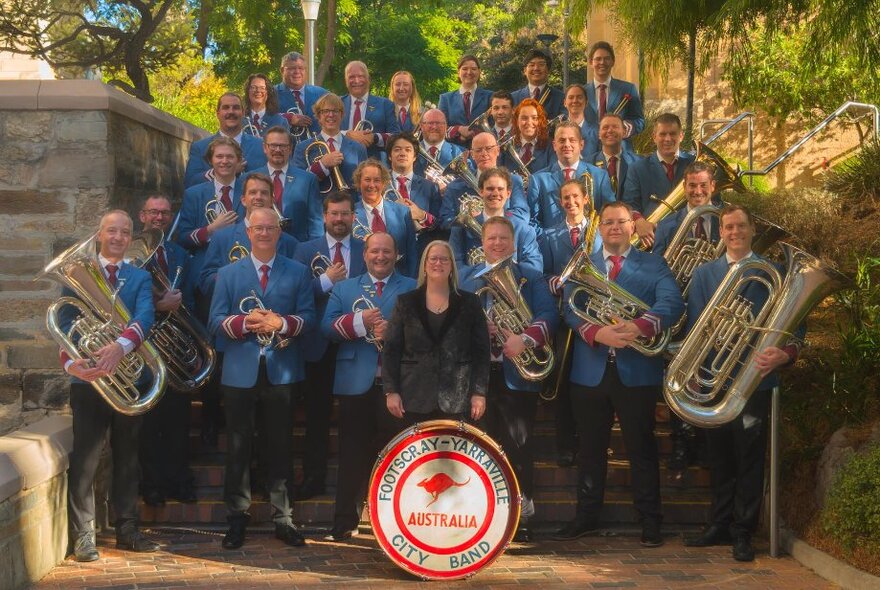 Footscray-Yarraville City Band, all wearing matching blue jackets and dark pants, posed with their instruments for a group photo outdoors on tiered steps, trees behind them.