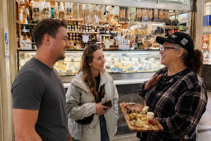 Two people in front of a market deli talk to a woman in a cap holding a tray of sample goods.