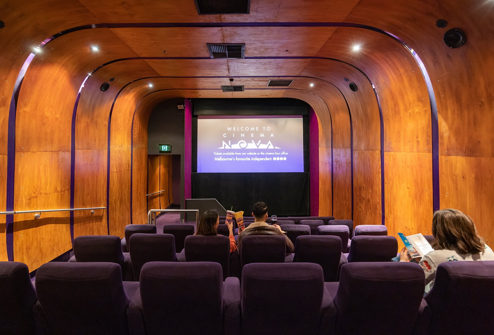 Three people sitting in a small movie theatre with curved timber walls and ceiling and purple seats.