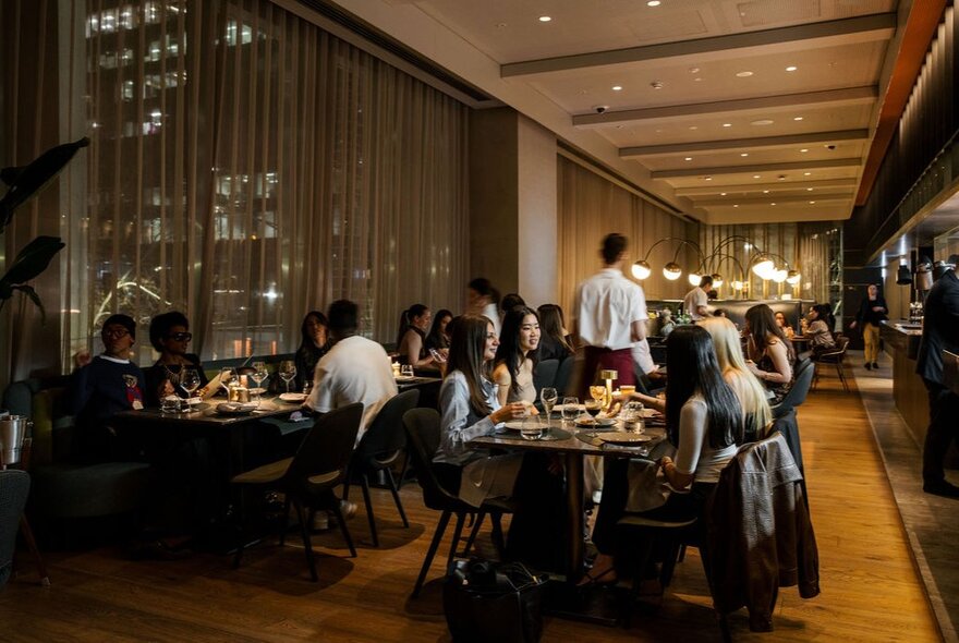 An upscale, dimly lit restaurant dining room with people sitting at tables next to large windows covered by sheer curtains.