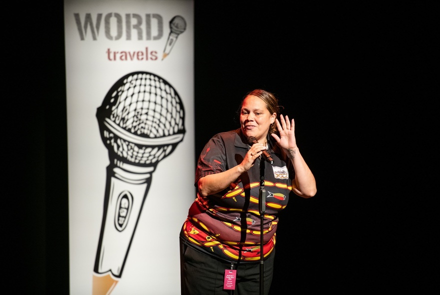 Performer on stage with hands raised, with a poster with the image of a microphone behind her.