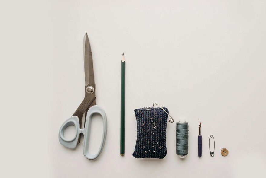 Sewing materials all lined up, including scissors, pencil, pin cushion, thread, needles and a safety pin. 