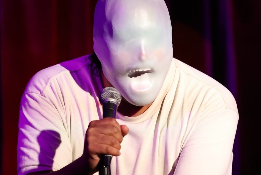 Comedian with a white rubber mask over his face, performing on a stage, holding a microphone in his hand.