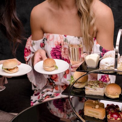 A woman in a floral strapless dress holding a small plate and champagne glass, seated behind a three-tiered cake stand with small delicacies.