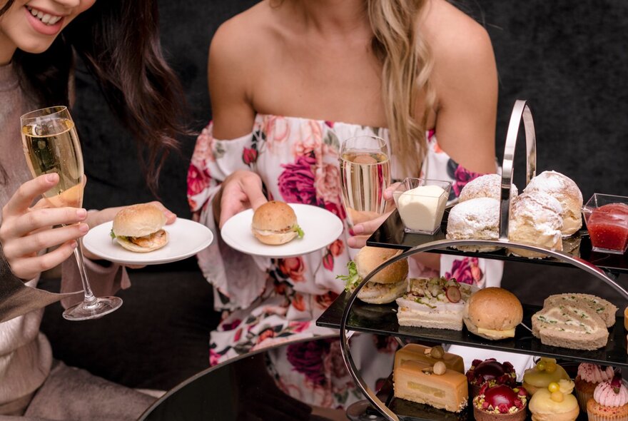 A woman in a floral strapless dress holding a small plate and champagne glass, seated behind a three-tiered cake stand with small delicacies.