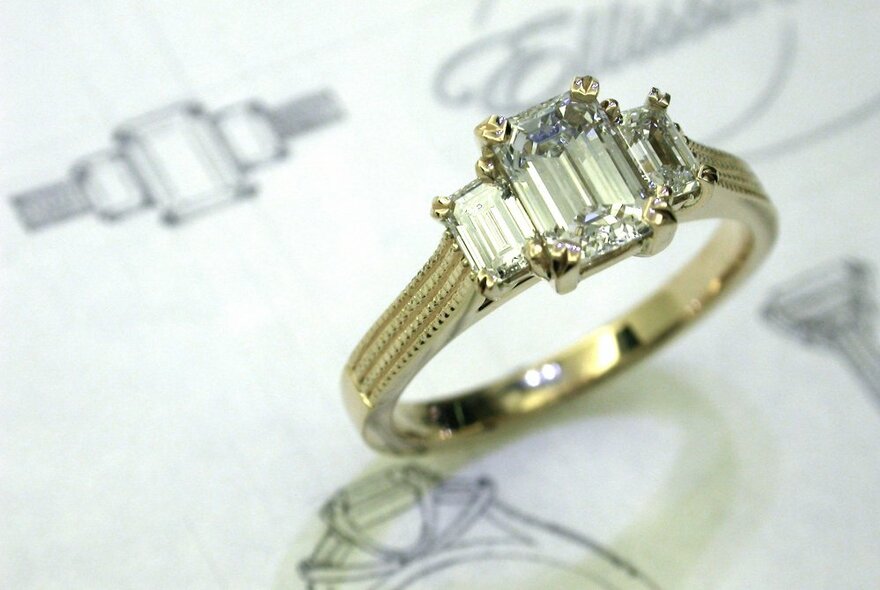 A gold engagement ring with three emerald cut diamonds, resting on a paper with its matching hand-drawn design.