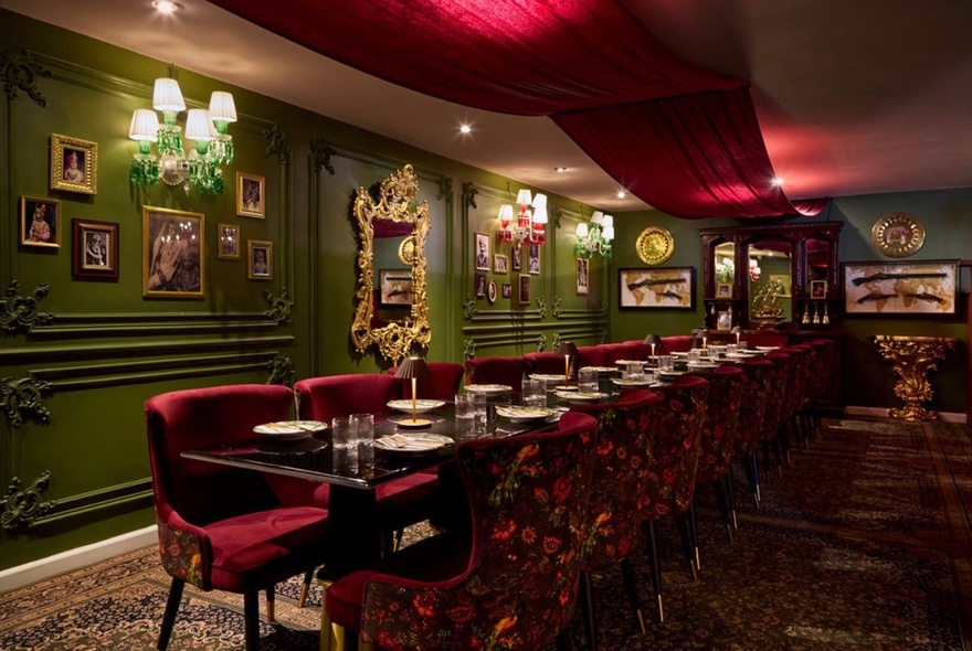 Indian restaurant banquet room with long table set for dinner with red velvet chairs, green walls decorated with paintings and lamps.