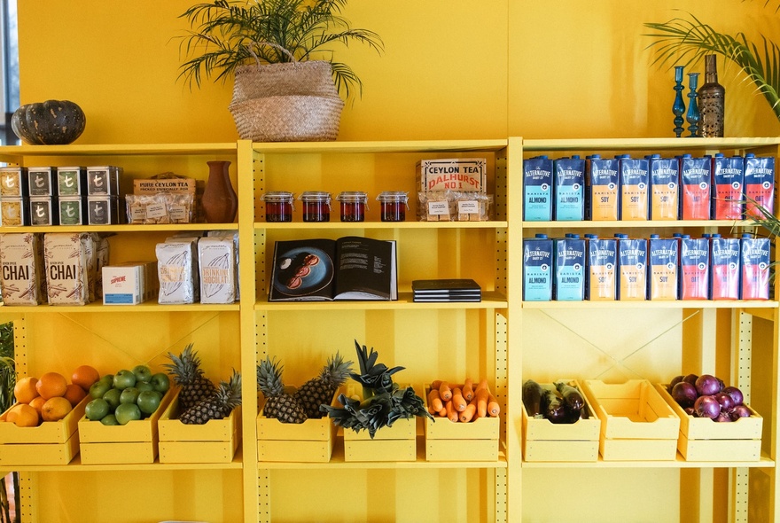 Two shelves of take-home Sri Lankan food and other pantry staples above a shelf of fresh fruit and vegetables; shelves and wall are painted yellow.