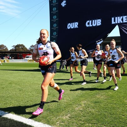 The Carlton AFLW team running through the cheering squad banner and onto the field.