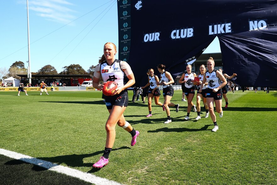 The Carlton AFLW team running through the cheering squad banner and onto the field.