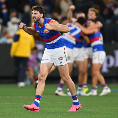 A huddle of Western Bulldogs AFL players celebrating on the field after having scored, with blurred fans in the stadium in the background.
