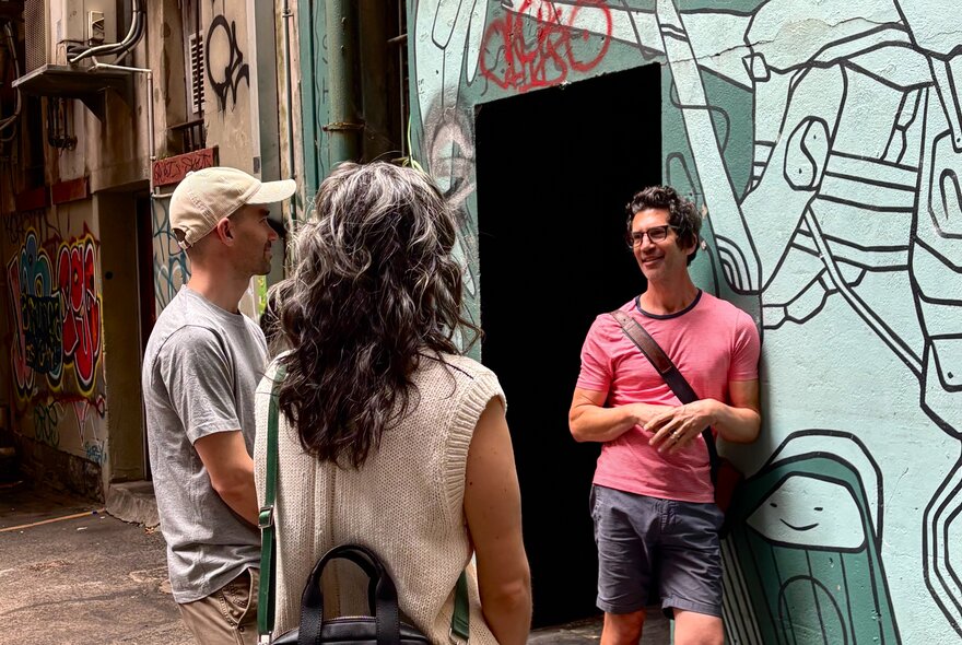 A tour guide wearing a pink t-shirt, talking to a couple on a walking tour about the mural art on the wall behind him.