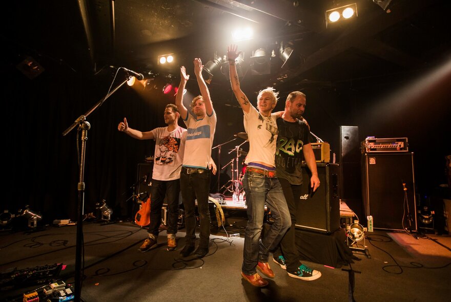 A still from the documentary film 'Jebediah: Are We OK?' shows all four band members on stage clapping and cheering after a concert. 