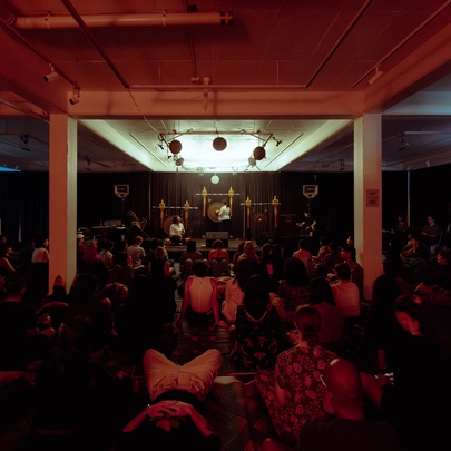 An indoor musical performance on a stage with large gong-like instruments under red lighting, with the audience seated on the floor.