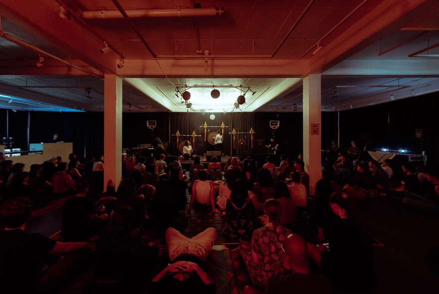 An indoor musical performance on a stage with large gong-like instruments under red lighting, with the audience seated on the floor.