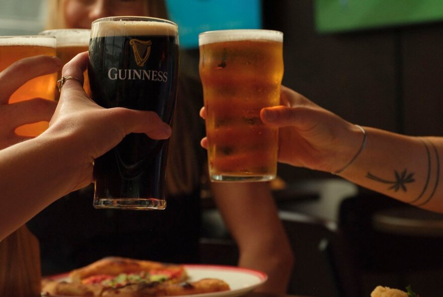 Two hands holding glasses of beer and bringing them together over a plate of food, in  a bar.
