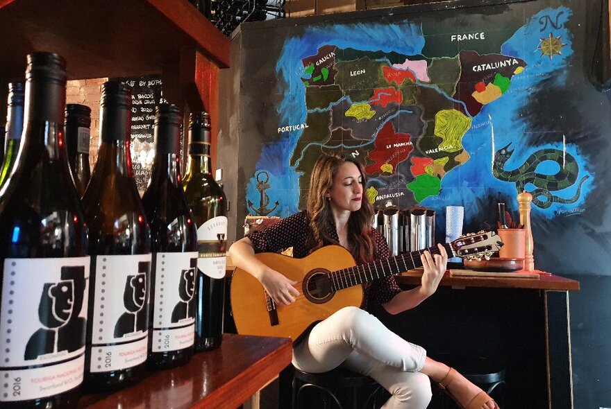 Laura de Elvira playing acoustic guitar indoors in a small restaurant with bottles of wine in a shelf. 