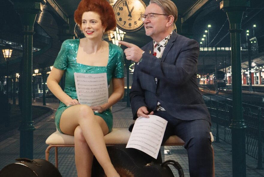 Man and woman seated on a bench in a dark venue with clock and seating, holding sheets of music.