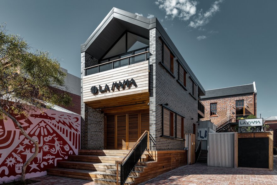The exterior of a black brick building with wooden steps and a red and pink mural next to it.