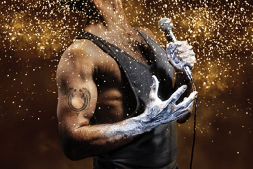 The well muscled torso of a man in a black singlet in profile view, holding a microphone in his hand, with flecks of gold dust showering around him from above.