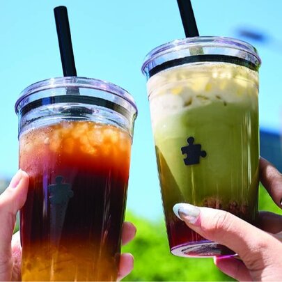 Two flavoured matcha or bubble drinks in clear plastic cups being held up by two hands against a blue sky.