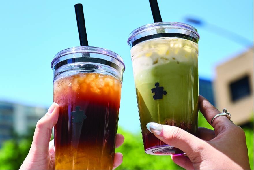 Two flavoured matcha or bubble drinks in clear plastic cups being held up by two hands against a blue sky.