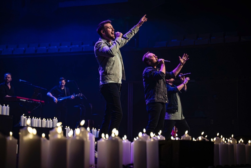 Three men on a candlelit stage, singing into microphones with arms outstretched.