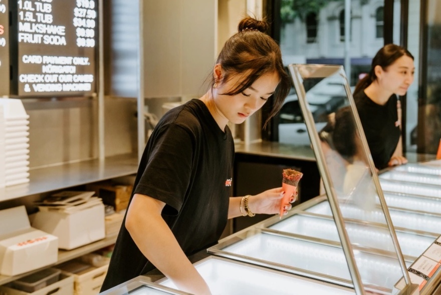 A woman scooping ice cream into a cone at a shop, with another woman serving in the background. 