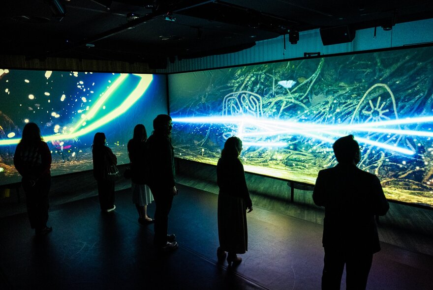 People in a darkened gallery space watching large projections with swirly, squiggly lines of light.