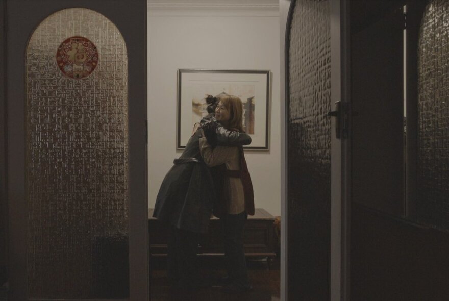Looking through an open door at two people hugging each other inside a house.
