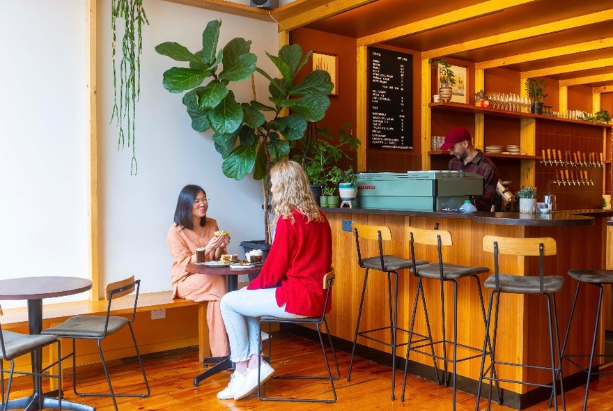 Two people sitting at a table to left of wooden bar and iron and wooden stools; large pot plant behind them.