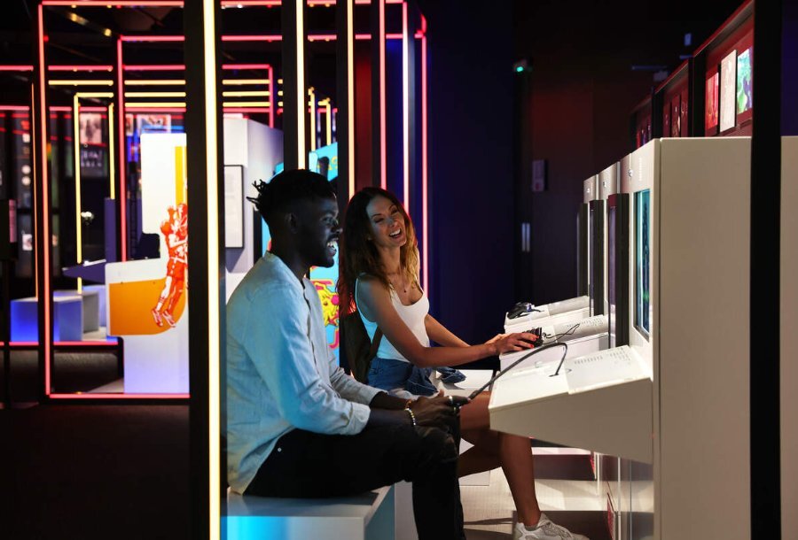 A man and woman playing an arcade game in a low-lit exhibit with neon lights.