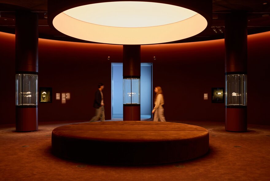 Two friends are in a gallery space with cabinets of jewellery there is a large circular velvet seat under a large circular light well 