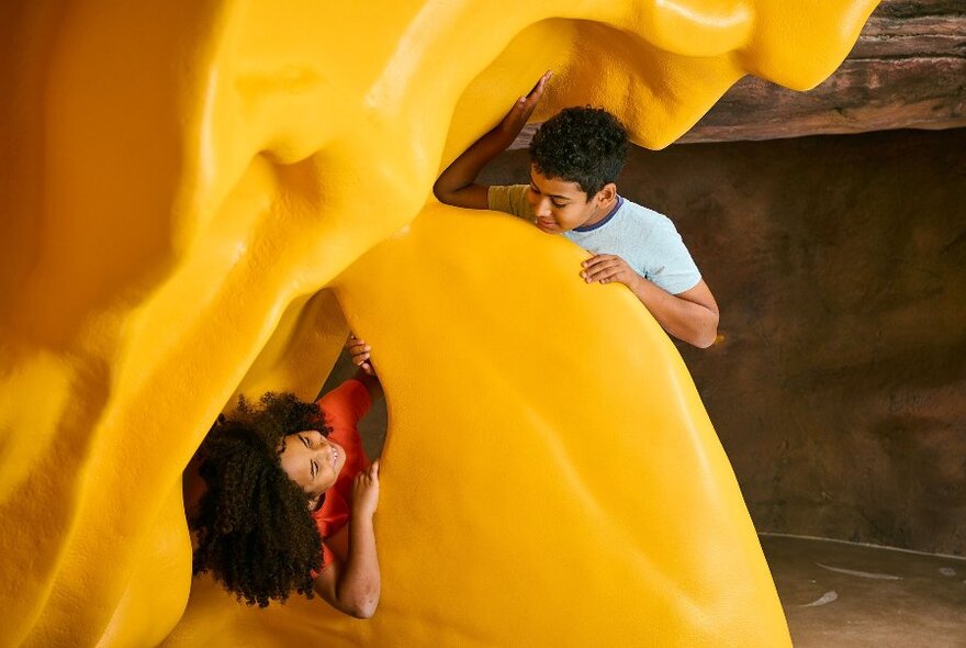 Two children looking at each while playing on a large, yellow sculpture, indoors.