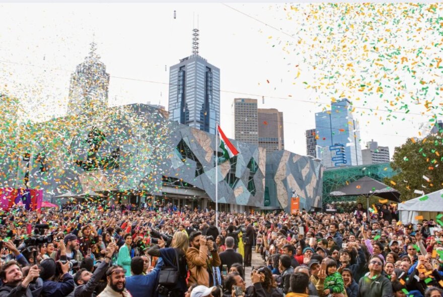 Orange, green and white bits of paper floating from the sky above a large crowd of people in the outdoor area at Fed Square, celebrating a flag raising ceremony.