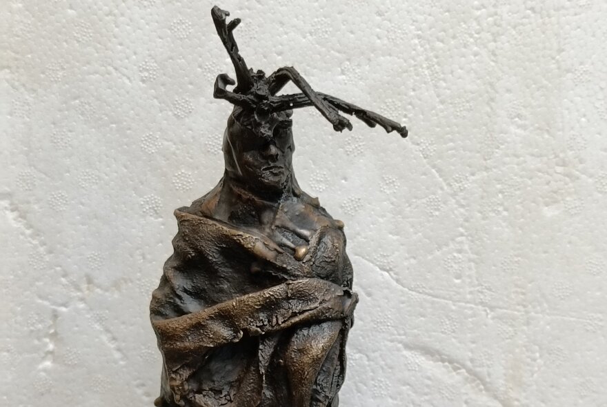 Bronze sculpture of a human figure with its torso  wrapped in fabric with three sticks protruding from its head.
