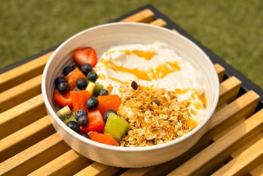 A healthy looking breakfast bowl of fruit, granola and yoghurt with honey, in bright sunlight on a wooden striped board on fake grass. 