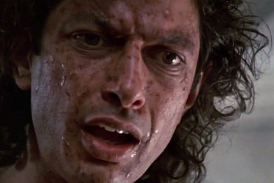 A still from a film showing the sweat-soaked face of a man with curly black hair as he looks at something in horror.