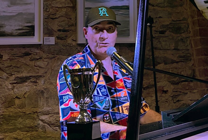 A man in a colourful shirt and baseball cap, seated at a grand piano and singing into a microphone.