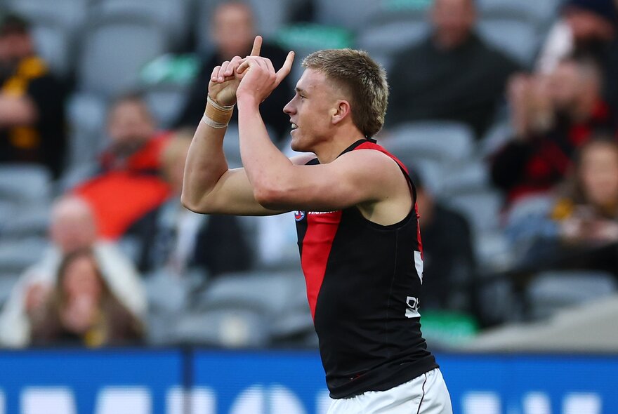 An Essendon AFL player smiling with his arms raised and fingers pointing upwards, and a blurred stadium crowd behind him.